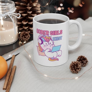May include: White ceramic mug with a pink, blue, and yellow unicorn wearing headphones and using a laptop. The text "CODER GIRLS EXIST" is printed on the mug.