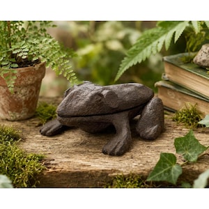 Pode incluir: Uma escultura de rã em ferro fundido castanho escuro com uma superfície texturizada. A rã está posicionada numa superfície de madeira com musgo e vegetação no fundo. A escultura é um elemento decorativo de jardim.