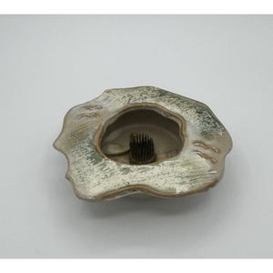 Puede incluir: Un ranero de cerámica con una forma orgánica texturizada y un pozo central. El ranero es principalmente de color verde apagado y beige, con un soporte de alfiler de metal oscuro en el centro. Los bordes tienen un diseño ondulado.