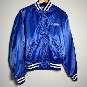 May include: A royal blue satin jacket with a button-up front, two pockets, and white and navy striped trim on the cuffs, collar, and waist. The jacket has the word "Paslode" embroidered on the chest.