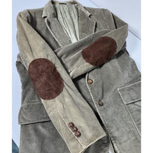 Puede incluir: Blazer de pana color beige con coderas de ante marrón. El blazer tiene solapas con muescas, dos bolsillos delanteros y tres botones en cada puño. Los botones son de color marrón oscuro.