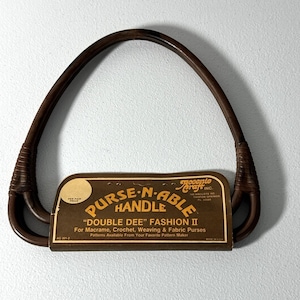 May include: A dark brown, D-shaped purse handle attached to a cardboard display card. The card features the text PURSE-N-ABLE HANDLE, DOUBLE DEE FASHION II, and Accento Craft Inc. The handle is designed for macrame, crochet, weaving, and fabric purses.