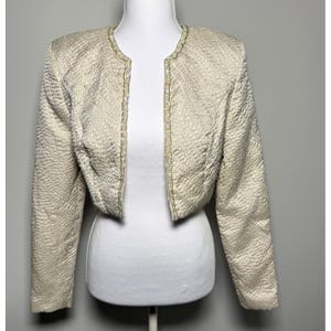 May include: Cream-colored, textured bolero jacket with a scalloped trim. The open-front jacket has long sleeves and is displayed on a white mannequin. The fabric has a subtle pattern, and the trim is a lighter shade of cream.