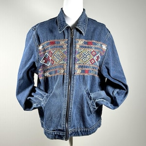 May include: A blue denim jacket with a full-length zipper and two front pockets. The jacket features colorful embroidered geometric patterns in red, yellow, and purple across the chest. The jacket is displayed on a mannequin.
