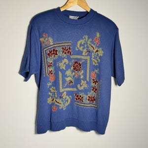 May include: A short-sleeved, periwinkle blue sweater with a crew neck. The sweater features a square floral design in the center, with burgundy, gold, and peach-colored flowers and leaves. The sweater is hanging on a wooden hanger.