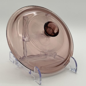 May include: A vintage, round, light purple glass lid with a matching knob. The lid features a radial design on the interior and is displayed on a clear plastic stand. This kitchenware item is likely for a casserole dish or pot.