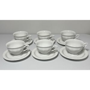 May include: Six white teacups and saucers are arranged in two rows. The cups have a rounded shape with a handle and sit on matching saucers. The saucers have a decorative edge. The set is made of porcelain.