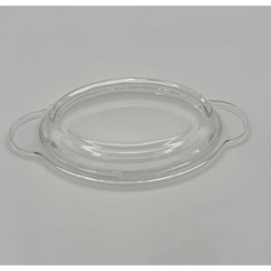 Puede incluir: Una tapa de cristal transparente de forma ovalada con una cúpula central elevada y dos asas laterales integradas. La tapa tiene una superficie lisa y un borde definido diseñado para encajar de forma segura en un recipiente o plato.