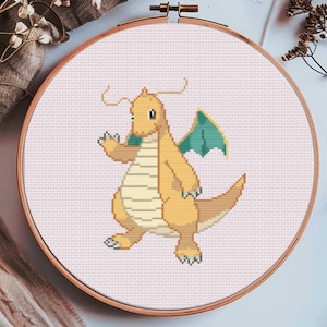 May include: A cross-stitch embroidery hoop featuring a pixelated Dragonite character. The design showcases the orange and yellow dragon with teal wings, set against a light pink background. The wooden hoop is visible, and the piece is surrounded by dried flowers.