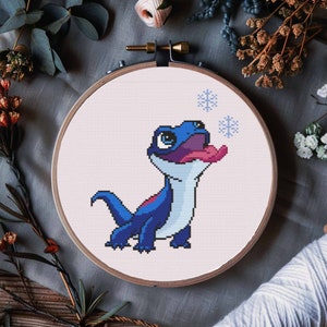 May include: Cross stitch pattern of a blue and purple lizard with a pink tongue, catching snowflakes. The pattern is in a round embroidery hoop.