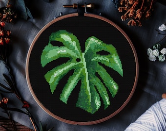 May include: A cross-stitch embroidery of a green monstera leaf on a black background, set in a wooden hoop. The image is surrounded by fabric, dried flowers, and a spool of white thread, creating a craft-themed aesthetic.