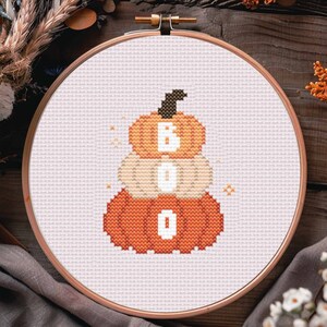 May include: A white embroidery hoop with a cross-stitch design of three orange pumpkins stacked on top of each other. The pumpkins spell out the word "BOO" in white.