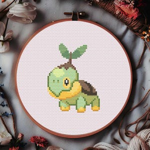 May include: A cross-stitch embroidery of a green and yellow turtle-like creature with a plant on its head, set in a wooden hoop. The design is on a white fabric background. The hoop is surrounded by flowers and yarn.