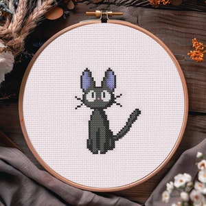May include: A black cat with purple ears and yellow eyes is embroidered on a white canvas in a wooden embroidery hoop.