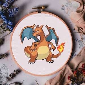 May include: A cross-stitch embroidery hoop featuring a pixelated image of a red and orange dragon with blue wings and a flame tail.