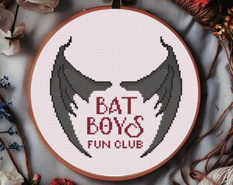 May include: A cross-stitch embroidery hoop featuring a design with dark gray bat wings and the text "BAT BOYS FUN CLUB" in burgundy. The embroidery is set against a light pink background, framed by a wooden hoop. The image is surrounded by flowers and yarn.