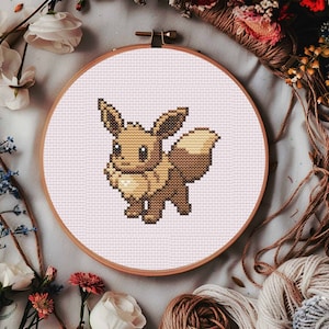 Könnte beinhalten: Eine braune und beigefarbene Kreuzstich-Stickerei eines Pokemon-Charakters, Eevee, auf rosa Hintergrund. Die Stickerei ist in einem Holzrahmen eingefasst.