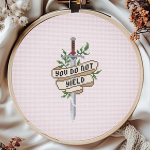 May include: A cross-stitch embroidery in a wooden hoop. The design features a sword piercing a banner with the words "YOU DO NOT YIELD" in black. Green leaves and red accents surround the sword and banner, set against a light pink background.