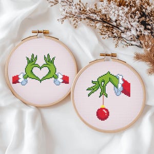 May include: Two cross-stitch hoops with Grinch-themed designs. One shows green hands forming a heart, the other a hand holding a red ornament. The hoops have wooden frames and are set against a light pink background.