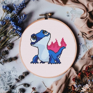 May include: A blue and pink cross-stitch embroidery of a cartoon lizard with a fire tail. The embroidery is framed in a wooden hoop.