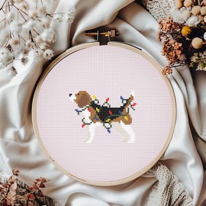 Può includere: Un telaio da ricamo a punto croce con un cane beagle decorato con luci natalizie. Il cane è marrone, nero e bianco, con luci colorate. Lo sfondo è rosa chiaro e il telaio è in legno.