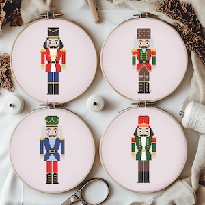 May include: Four cross-stitch embroidery hoops displaying nutcracker soldier designs. Each nutcracker wears a different uniform, with red, green, blue, and brown colours. The hoops are set against a light pink background, with wooden hoops and white thread.