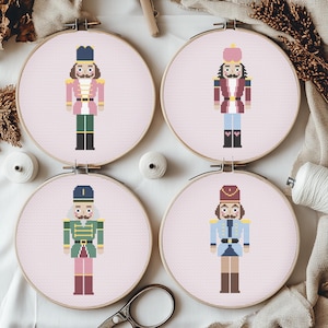 Könnte beinhalten: Vier Kreuzstich-Stickrahmen mit pixeligen Nussknacker-Soldaten-Designs. Jeder Nussknacker hat ein anderes Uniformfarbschema, darunter Rosa, Grün, Rot und Blau. Der Hintergrund ist hellrosa, und die Reifen werden auf einer weißen Oberfläche angezeigt.