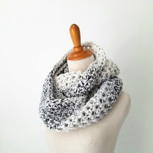 May include: A white and gray ombre infinity scarf made of crocheted yarn. The scarf is displayed on a white mannequin.