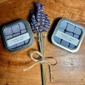 May include: Two small silver tins filled with lavender and lemon scented glitter cubes. The tins have labels that read "Lavender + Lemon Glitter Cubes". A bouquet of dried lavender is in the center of the image.