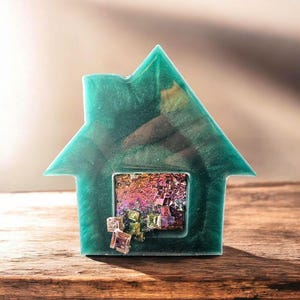 Puede incluir: Un pisapapeles de resina de color verde azulado en forma de casa con una ventana que revela un cristal iridiscente de colores en su interior.
