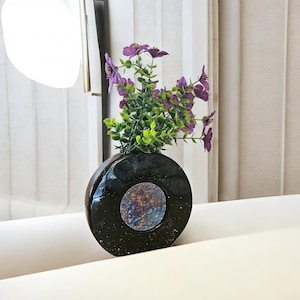 May include: A circular black vase with a glossy finish and white speckles, featuring a central circular inset with a textured, multi-coloured metallic finish. The vase holds a bouquet of artificial purple flowers with green leaves.