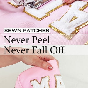 May include: Close-up of a pink fabric pouch with white letters that spell "MA" outlined in gold glitter. The letters are sewn on and have a fluffy texture. The text "SEWN PATCHES Never Peel Never Fall Off" is visible in the background.