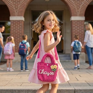 May include: A young girl waves while carrying a pink corduroy handbag with the name "Chayse" embroidered on it. The bag features a flower embellishment and a matching shoulder strap. She is wearing a pink dress and is in front of a brick building.