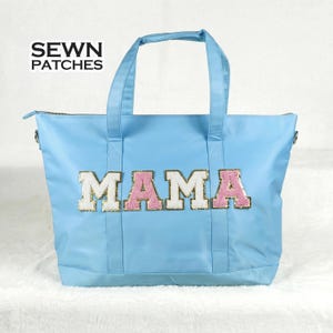 Puede incluir: Bolso tote de color azul claro con la palabra "MAMA" aplicada en la parte delantera con letras blancas y rosas con lentejuelas. El bolso tiene dos asas y un cierre de cremallera. El texto "SEWN PATCHES" está en la esquina superior izquierda.
