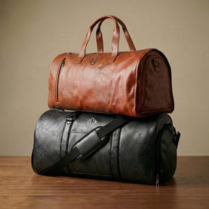 Puede incluir: Una bolsa de viaje de cuero marr&oacute;n con el nombre SIMON grabado en el frente se apoya sobre una bolsa de viaje de cuero negro con el monograma EGM. Ambas bolsas tienen dos asas superiores, cremalleras laterales y una correa para el hombro.