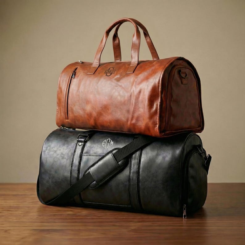 Puede incluir: Una bolsa de viaje de cuero marr&oacute;n con el nombre SIMON grabado en el frente se apoya sobre una bolsa de viaje de cuero negro con el monograma EGM. Ambas bolsas tienen dos asas superiores, cremalleras laterales y una correa para el hombro.