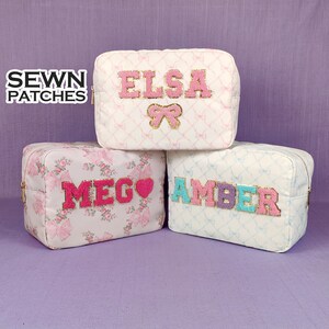 Puede incluir: Tres bolsas de cosméticos de tela blanca con letras bordadas en rosa, azul y dorado. Las bolsas están etiquetadas como "Elsa", "Meg" y "Amber".