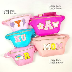 May include: Five pink, blue and light pink fanny packs with different letters and designs. The fanny packs are arranged in a semi-circle. The letters on the fanny packs are Y, A, KU, MOM and R. The designs on the fanny packs are a heart, a flower, a lightning bolt, a butterfly and a rainbow.