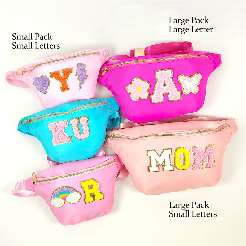 May include: Five pink, blue and light pink fanny packs with different letters and designs. The fanny packs are arranged in a semi-circle. The letters on the fanny packs are Y, A, KU, MOM and R. The designs on the fanny packs are a heart, a flower, a lightning bolt, a butterfly and a rainbow.