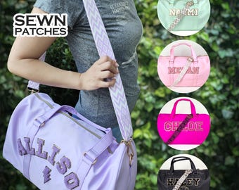 Puede incluir: Una bolsa de deporte de color morado con una cremallera dorada y un nombre bordado en oro "Allison" en la parte delantera. La bolsa tiene una correa a rayas blanca y morada. La imagen también muestra otras cuatro bolsas de deporte con diferentes nombres y colores.