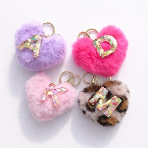 May include: Four heart-shaped keychains with gold-colored hardware. The keychains are made of fluffy material in purple, pink, and leopard print. Each keychain has a letter with glitter accents: A, D, K, and M.