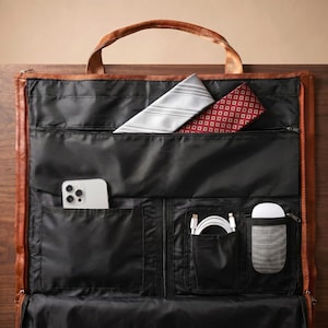 Puede incluir: Una funda para ropa de cuero marr&oacute;n con un interior negro y m&uacute;ltiples compartimentos de almacenamiento. La bolsa contiene un tel&eacute;fono inteligente blanco, un cable de carga blanco, un rat&oacute;n de ordenador blanco, una corbata gris a rayas y una corbata roja estampada.