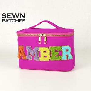 May include: A bright pink cosmetic bag with a gold zip and handle. The name "AMBER" is displayed in colourful, textured letters. The bag features the text "SEWN PATCHES" in black.