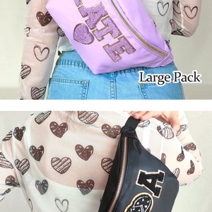 May include: Two fanny packs, one purple with the word "KAFE" in gold glitter letters and one black with a gold "A" and a heart design. Both fanny packs have gold zippers and are worn by models wearing a white shirt with brown hearts and blue jeans.