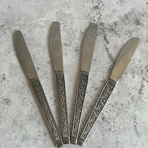 Peut inclure: Quatre couteaux à beurre argentés avec des motifs floraux ornés sur les manches. Les lames sont lisses et incurvées. Les couteaux sont disposés sur une surface grise et texturée, évoquant un style vintage ou antique.