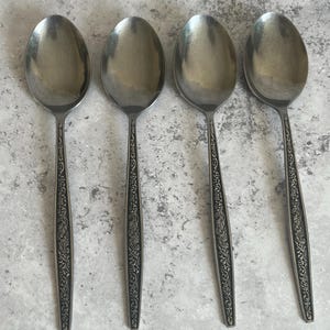 Può includere: Quattro cucchiai in acciaio inossidabile con coppette ovali e decorazioni floreali ornate sui manici. I cucchiai sono disposti in fila, mostrando le loro superfici lucide e riflettenti. Un classico set di posate.
