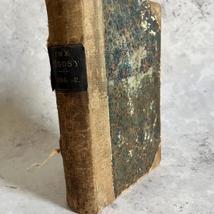 Könnte beinhalten: Ein antikes Buch mit dem Titel "The Argosy" mit einem abgenutzten, strukturierten Einband. Der Buchrücken ist beige mit einem schwarzen Etikett mit dem Titel und "1894. 2." Das Cover ist eine Mischung aus Beige und Dunkelgrün mit einem abgenutzten Aussehen.