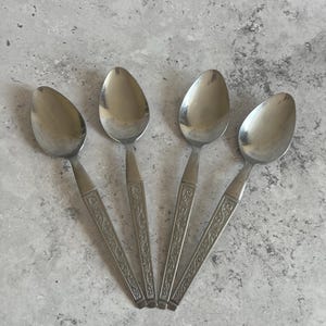 May include: Four stainless steel spoons arranged in a fan shape. Each spoon has an oval bowl and a handle with an engraved floral design. The spoons are set against a mottled grey background.