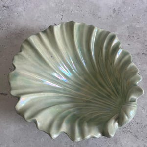 May include: A pale green, iridescent ceramic seashell-shaped dish. The dish has a ruffled edge and a textured, fan-like pattern radiating from the centre. The surface reflects a subtle rainbow sheen.