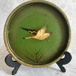 May include: A round, green tray with a gold rim and a painted image of a bird in flight. The bird is white with black wings and is flying over a field of green grass.